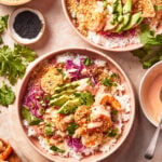 Two bowls of rice topped with shrimp, sliced avocado, shredded cabbage, green onions, cilantro, sauce, and sesame seeds, surrounded by ingredients like black sesame seeds and fresh cilantro on a beige surface.