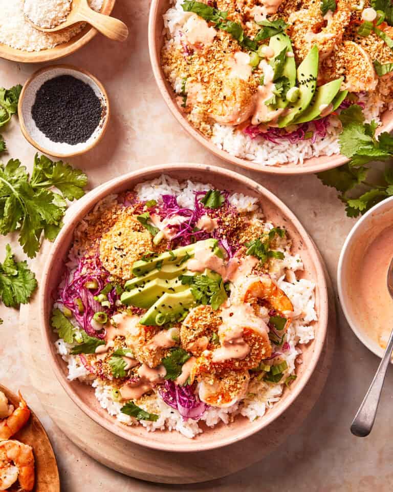 Two bowls of rice topped with shrimp, sliced avocado, shredded cabbage, green onions, cilantro, sauce, and sesame seeds, surrounded by ingredients like black sesame seeds and fresh cilantro on a beige surface.