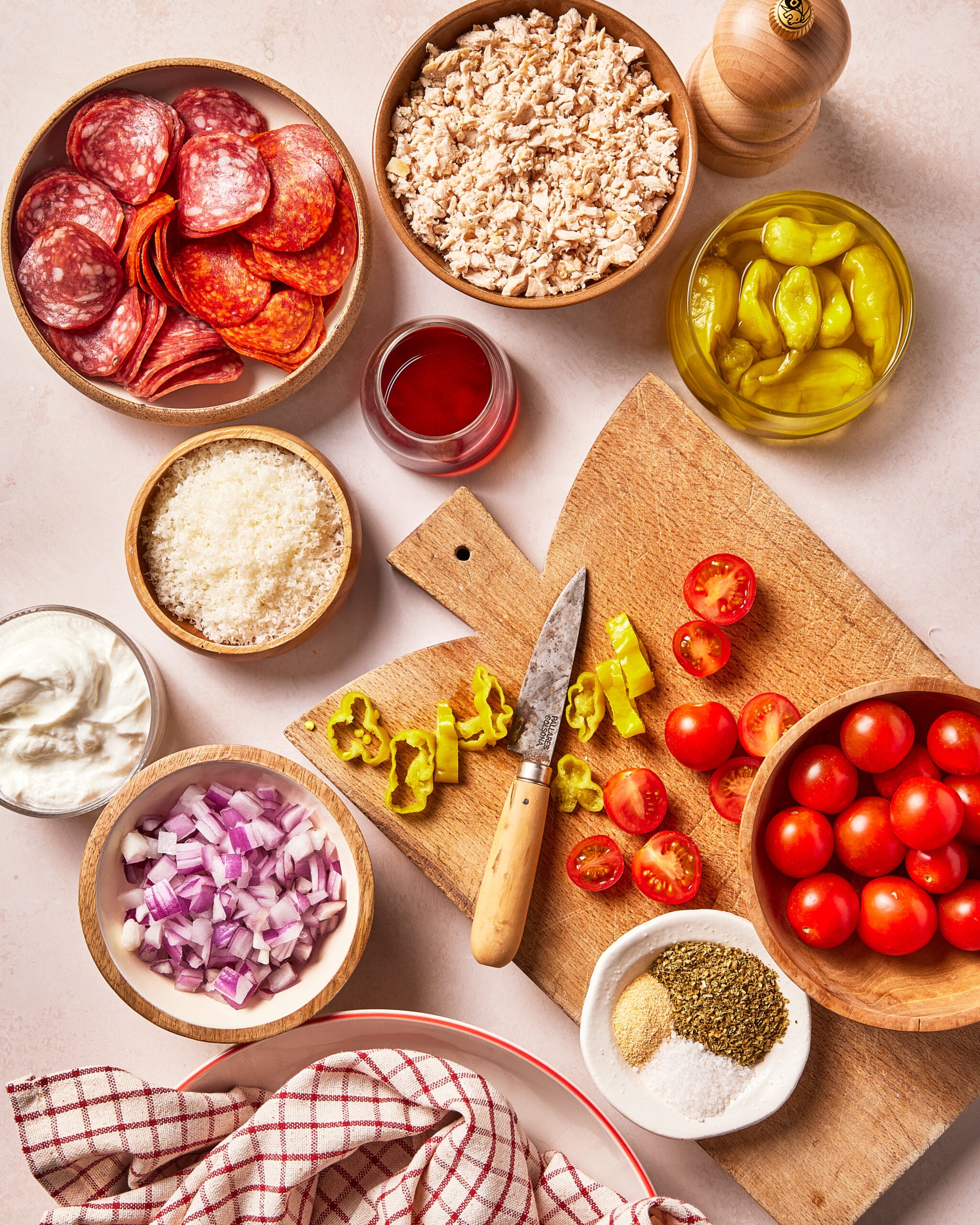 Ingredients for a salad laid out on a table, including bowls of sliced salami, shredded chicken, grated cheese, diced red onions, cherry tomatoes, pepperoncini, spices, dressing, and a wooden cutting board with a knife.