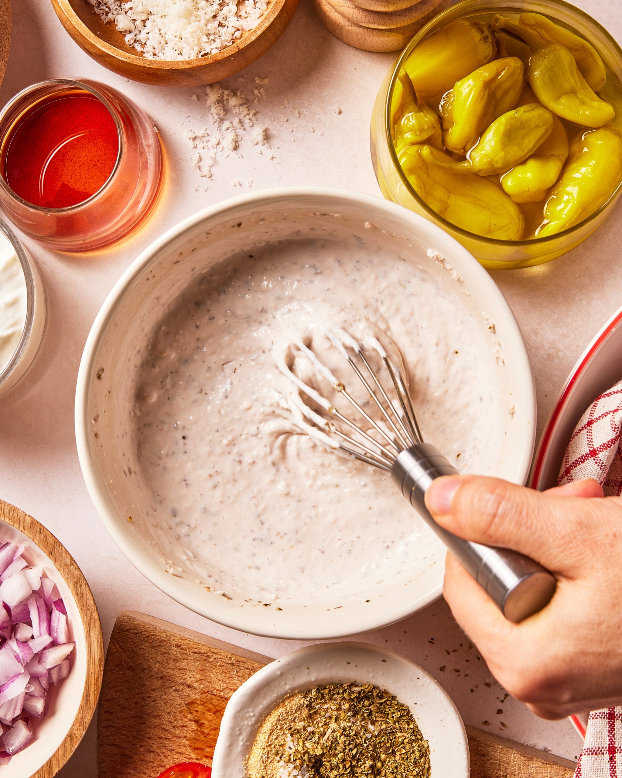 A hand whisks a creamy dressing in a white bowl, surrounded by small bowls containing diced red onion, grated cheese, dried herbs, a glass of red liquid, and a jar of pickled peppers.