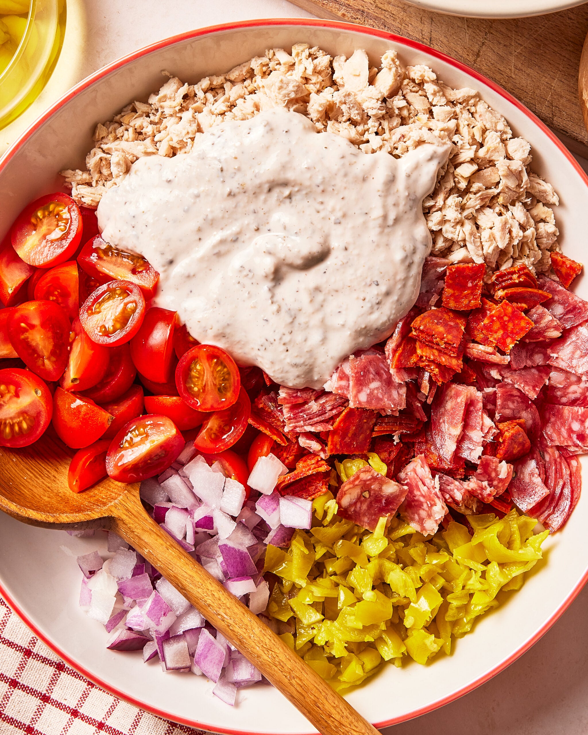 A salad bowl containing tuna, halved cherry tomatoes, chopped red onion, sliced pepperoni, and pepperoncini, topped with a dollop of creamy dressing and a wooden spoon.