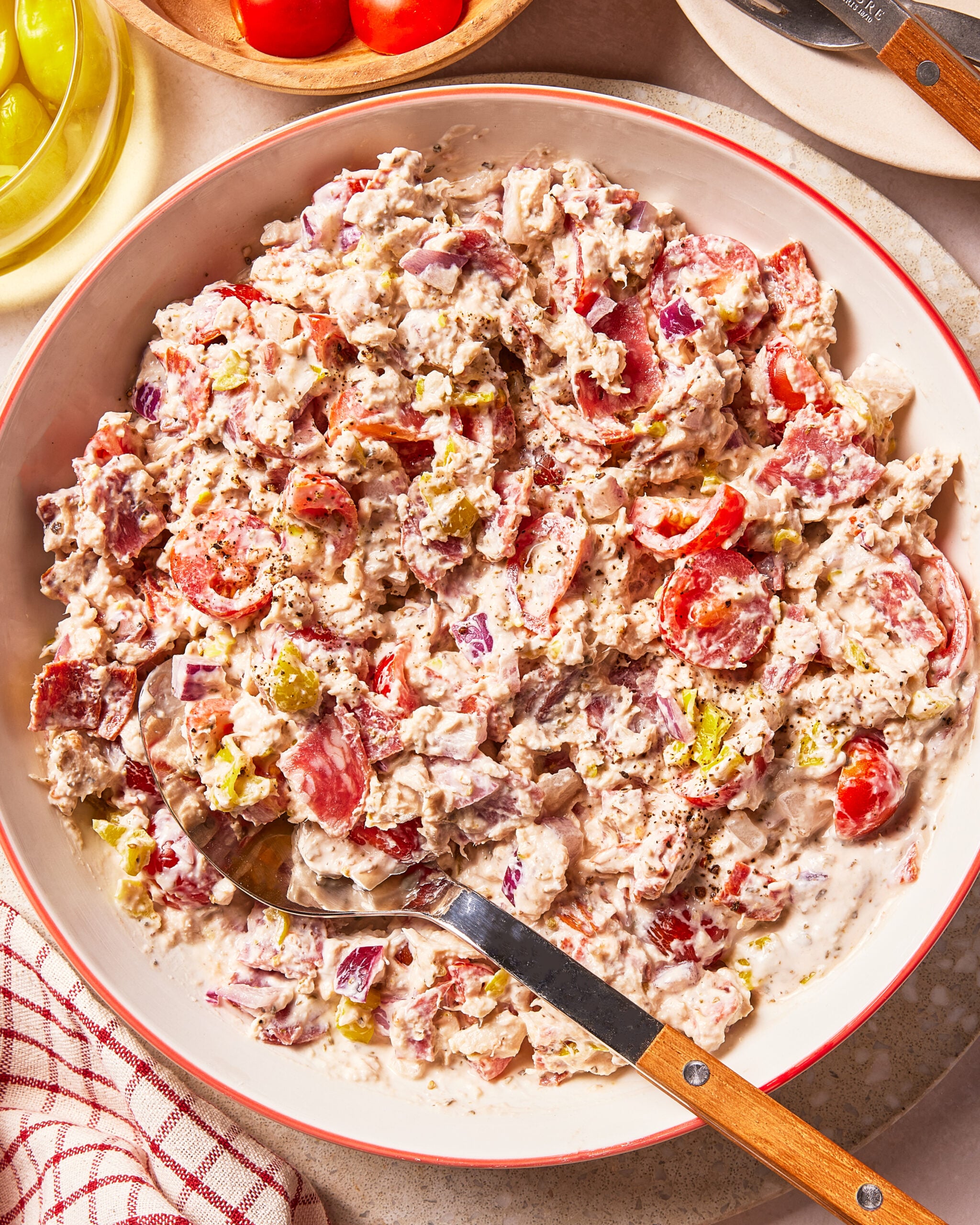 A bowl of creamy pasta salad containing salami, cherry tomatoes, and red onion, with a serving spoon resting inside.