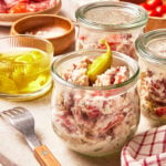 Glass jars filled with creamy chicken salad containing chunks of tomato and topped with a whole pepperoncini, next to a fork and bowls of ingredients.