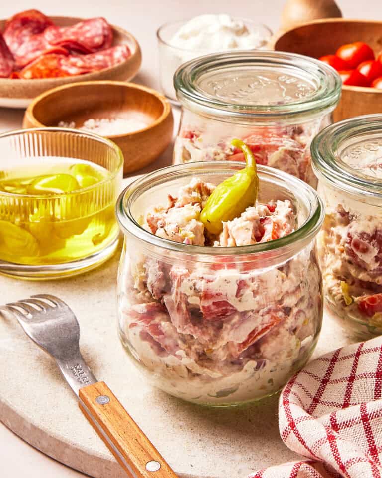 Glass jars filled with creamy chicken salad containing chunks of tomato and topped with a whole pepperoncini, next to a fork and bowls of ingredients.