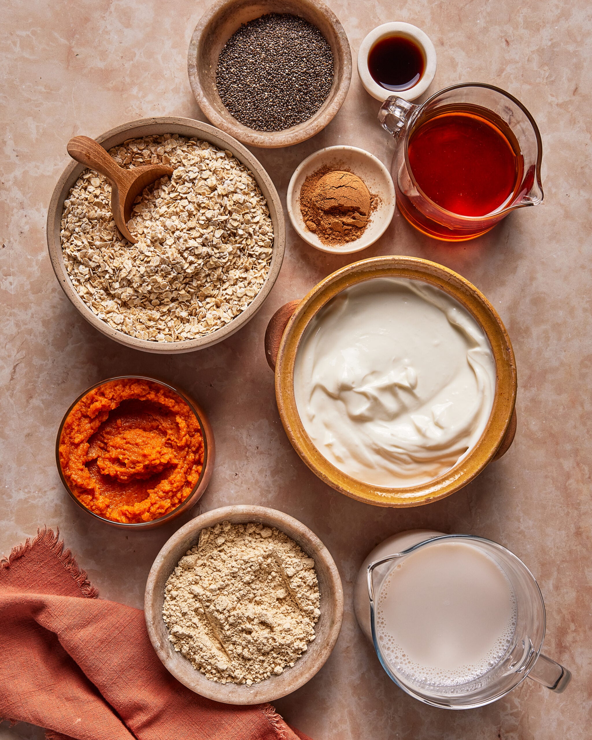 Overhead view of bowls containing oats, chia seeds, cinnamon, pumpkin purée, protein powder, yogurt, maple syrup, and milk.
