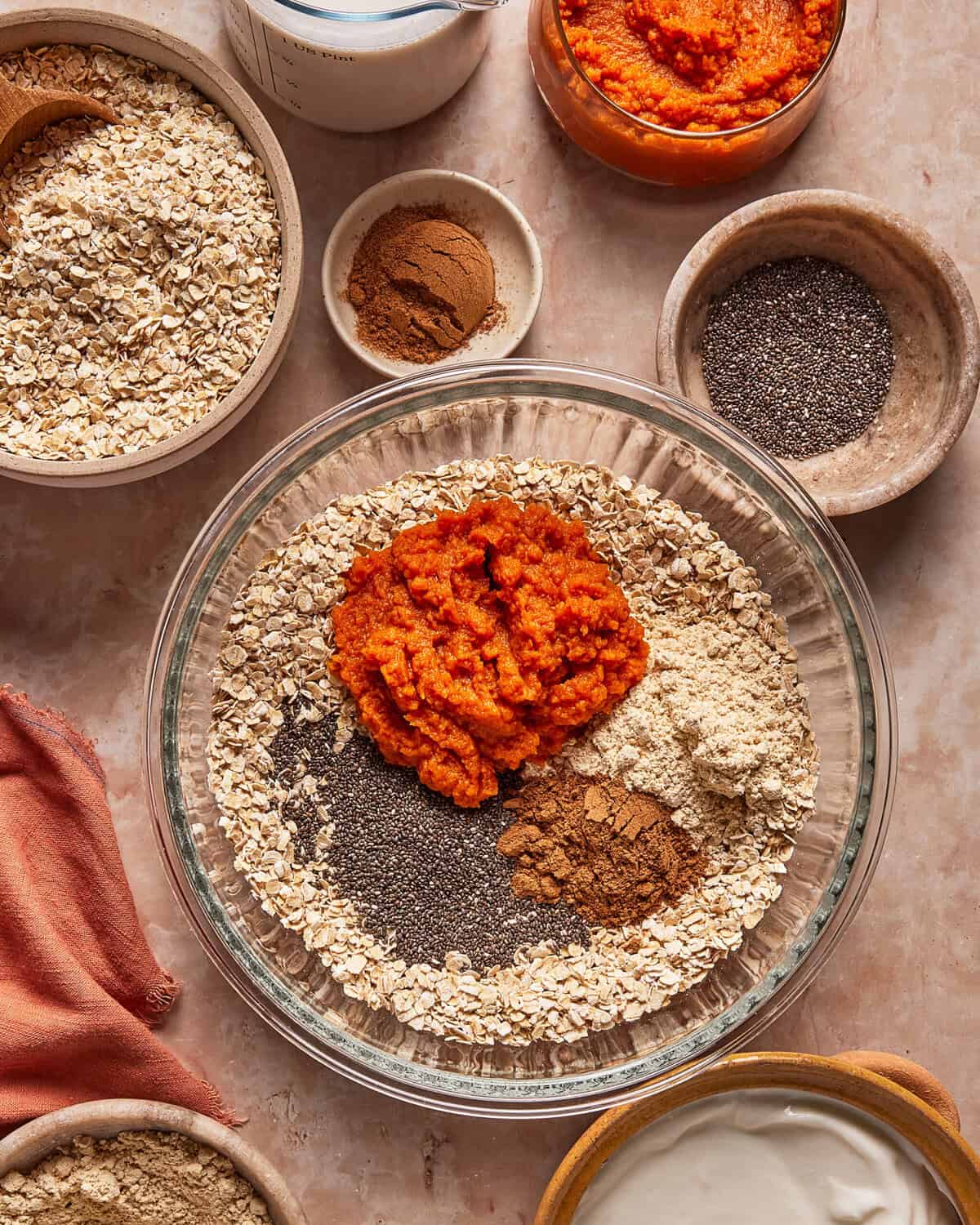 A large glass bowl contains rolled oats, chia seeds, pumpkin purée, and spices, surrounded by individual bowls of the same ingredients and a measuring cup.