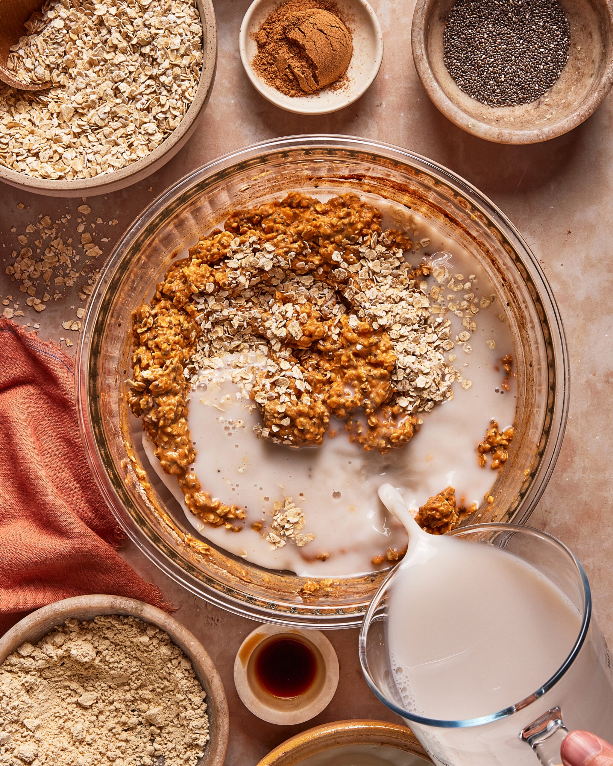 Milk being poured into a glass mixing bowl containing oats and a spiced mixture, surrounded by small bowls of ingredients including rolled oats, cinnamon, chia seeds, and protein powder.