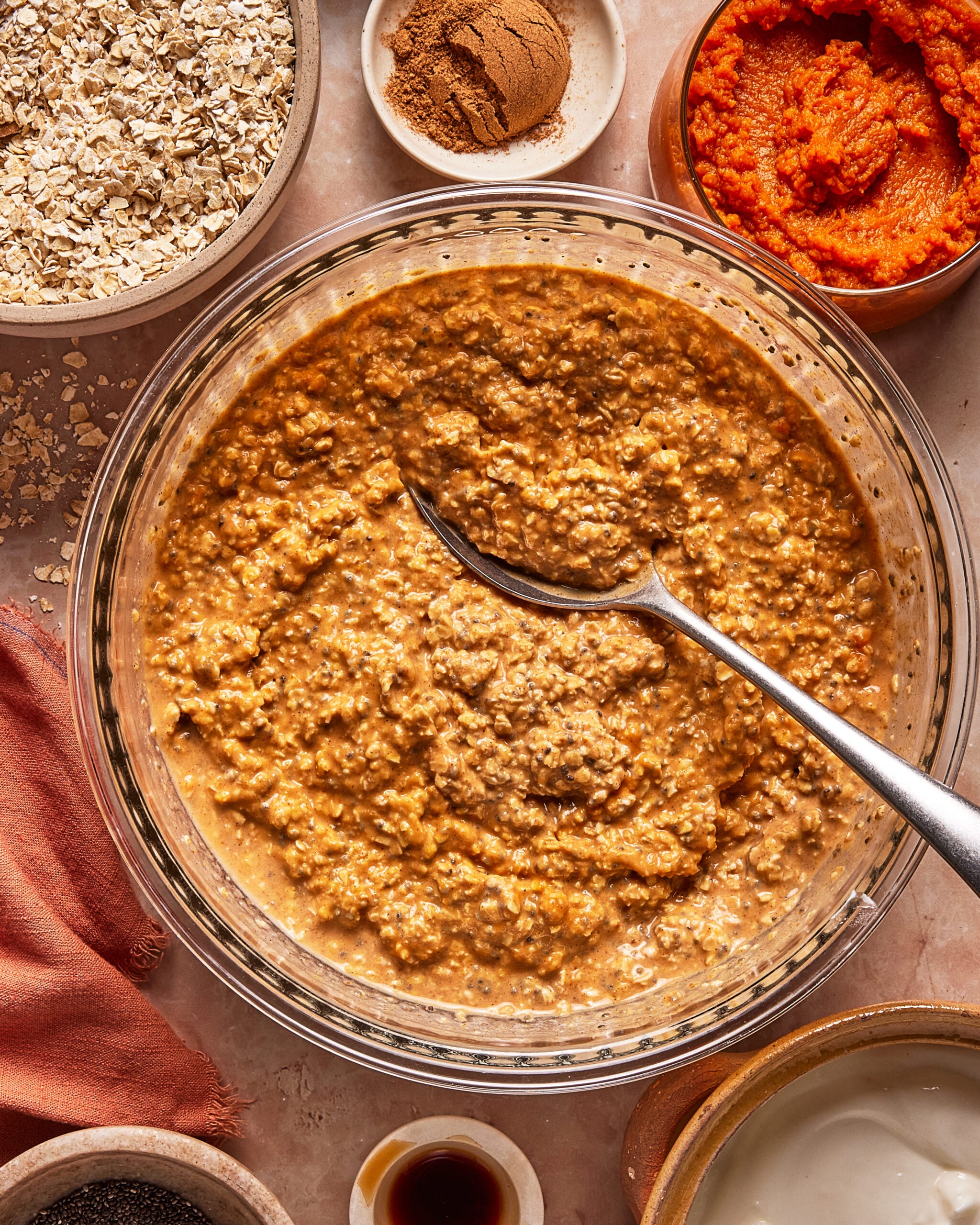 A glass bowl filled with pumpkin-colored oatmeal and a metal spoon, surrounded by bowls of raw oats, pumpkin puree, spices, and other ingredients.