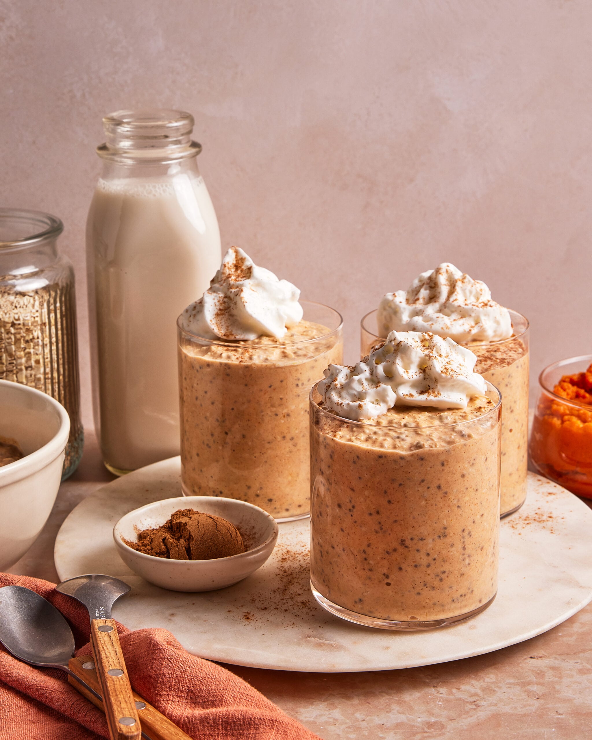 Three glasses of pumpkin overnight oats topped with whipped cream and cinnamon on a marble serving board, with a bottle of milk and bowls of ingredients nearby.