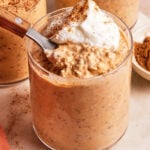 A glass filled with pumpkin overnight oats topped with whipped cream and a sprinkle of cinnamon, shown with a spoon resting inside.