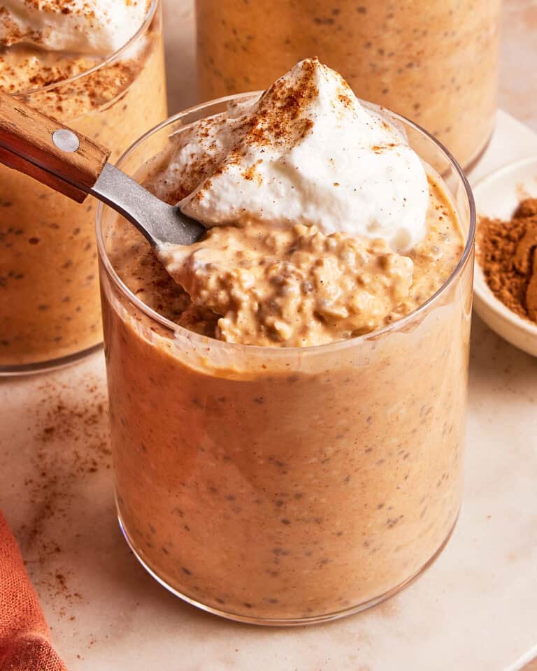 A glass filled with pumpkin overnight oats topped with whipped cream and a sprinkle of cinnamon, shown with a spoon resting inside.