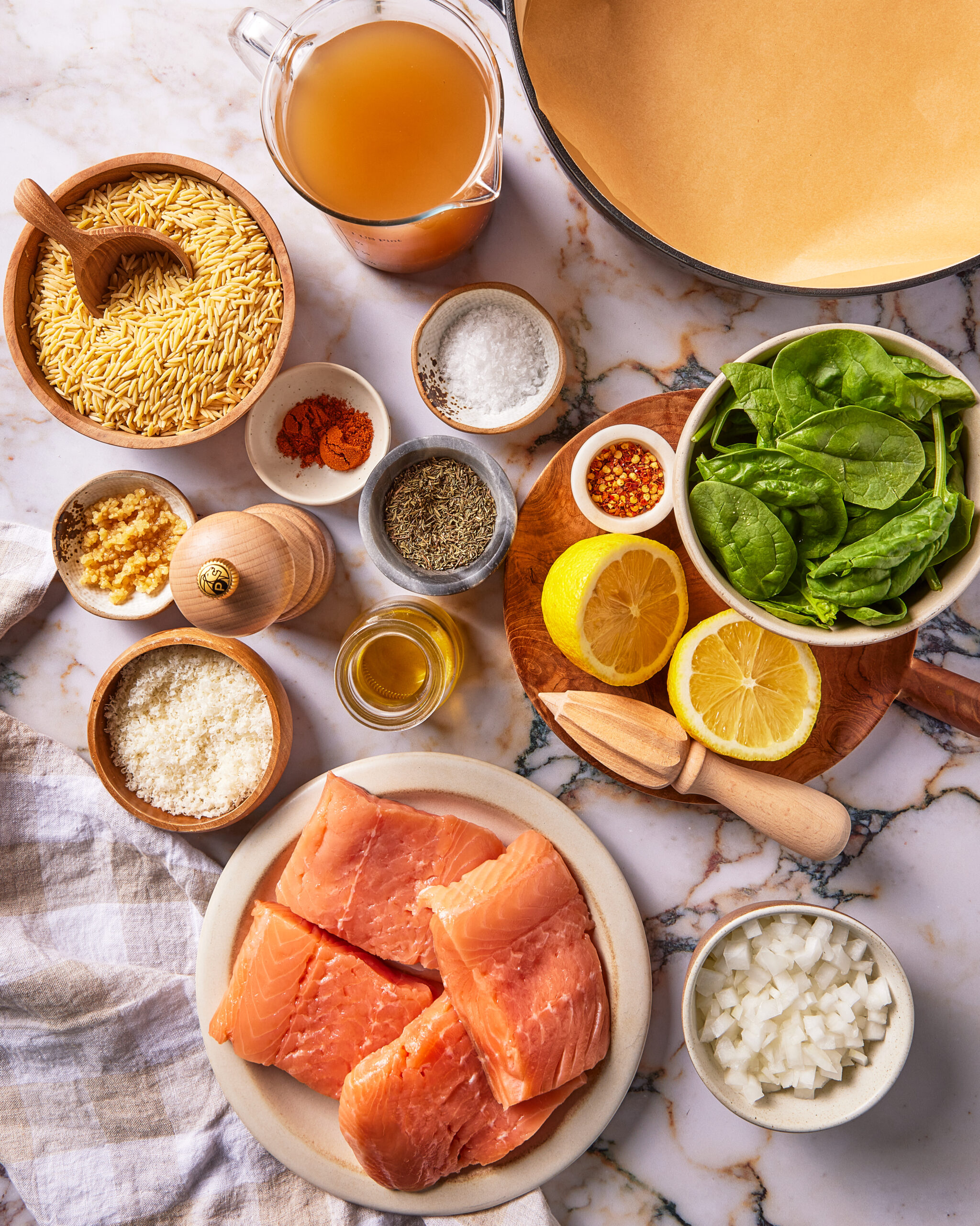 Ingredients for a salmon dish, including raw salmon fillets, orzo, spinach, lemon, broth, onion, garlic, spices, and a wooden citrus reamer on a marble surface.