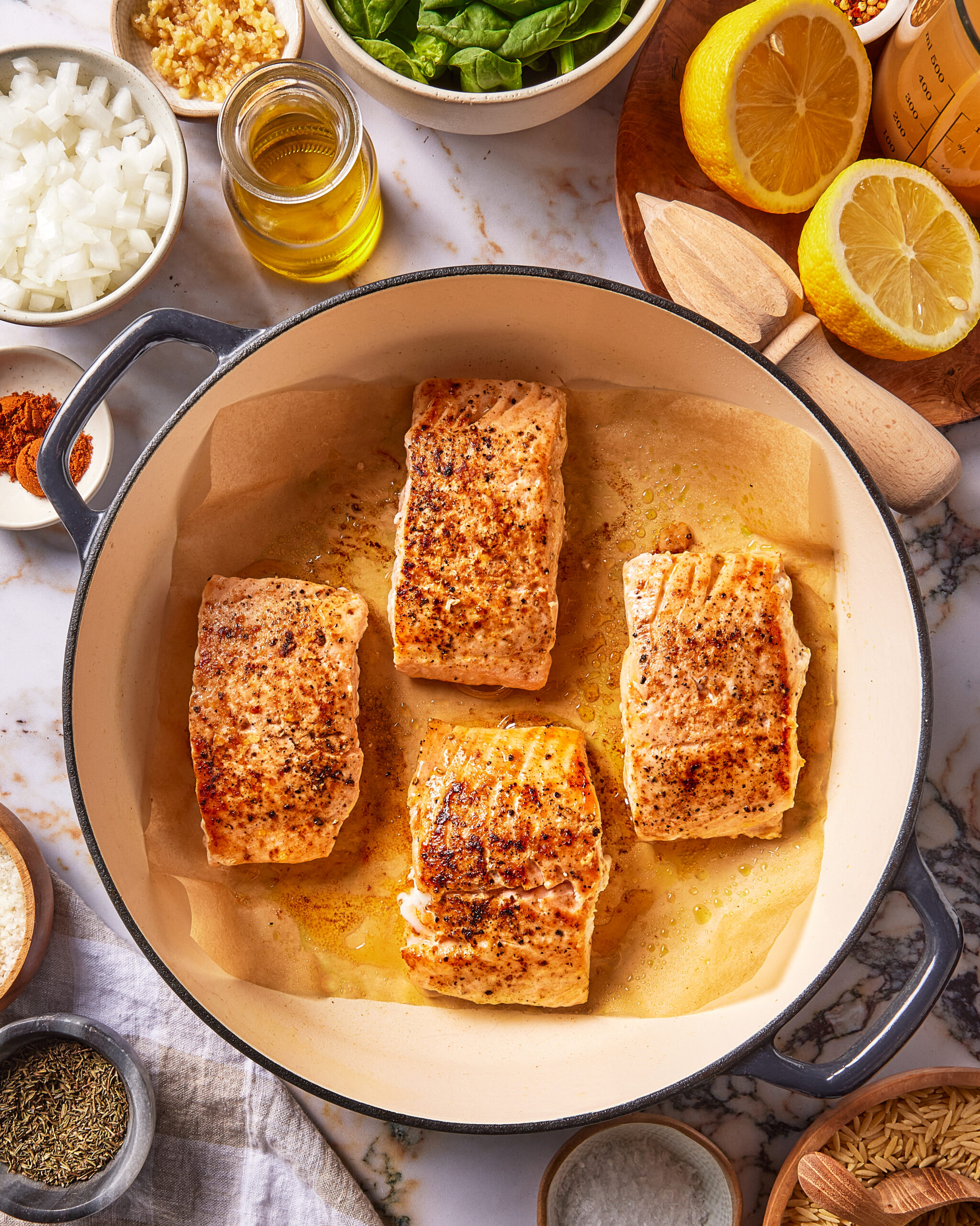 Four seasoned salmon fillets searing in a parchment-lined cast iron pan, surrounded by bowls of chopped onion, spinach, oil, and lemon halves.