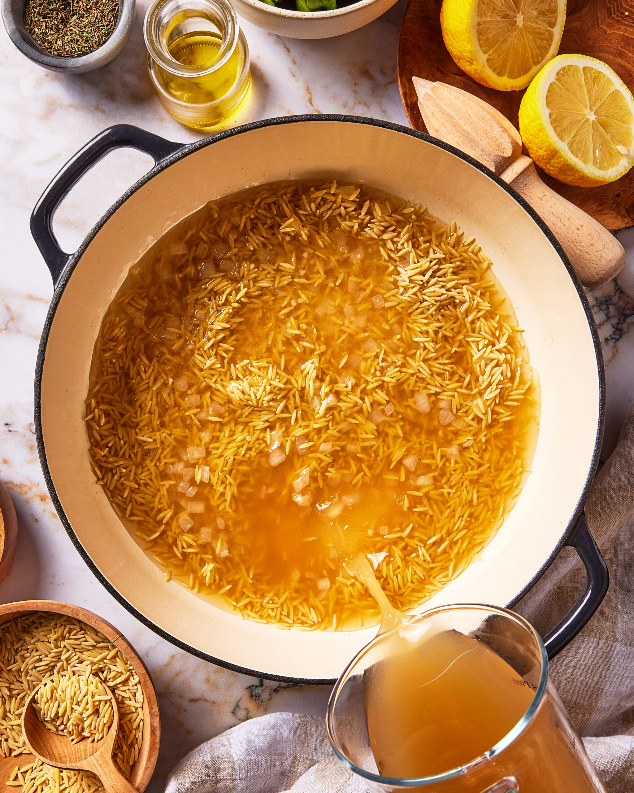 A liquid being poured from a glass measuring cup into a large pot containing orzo pasta and broth, surrounded by bowls of dried herbs, olive oil, and lemon halves.