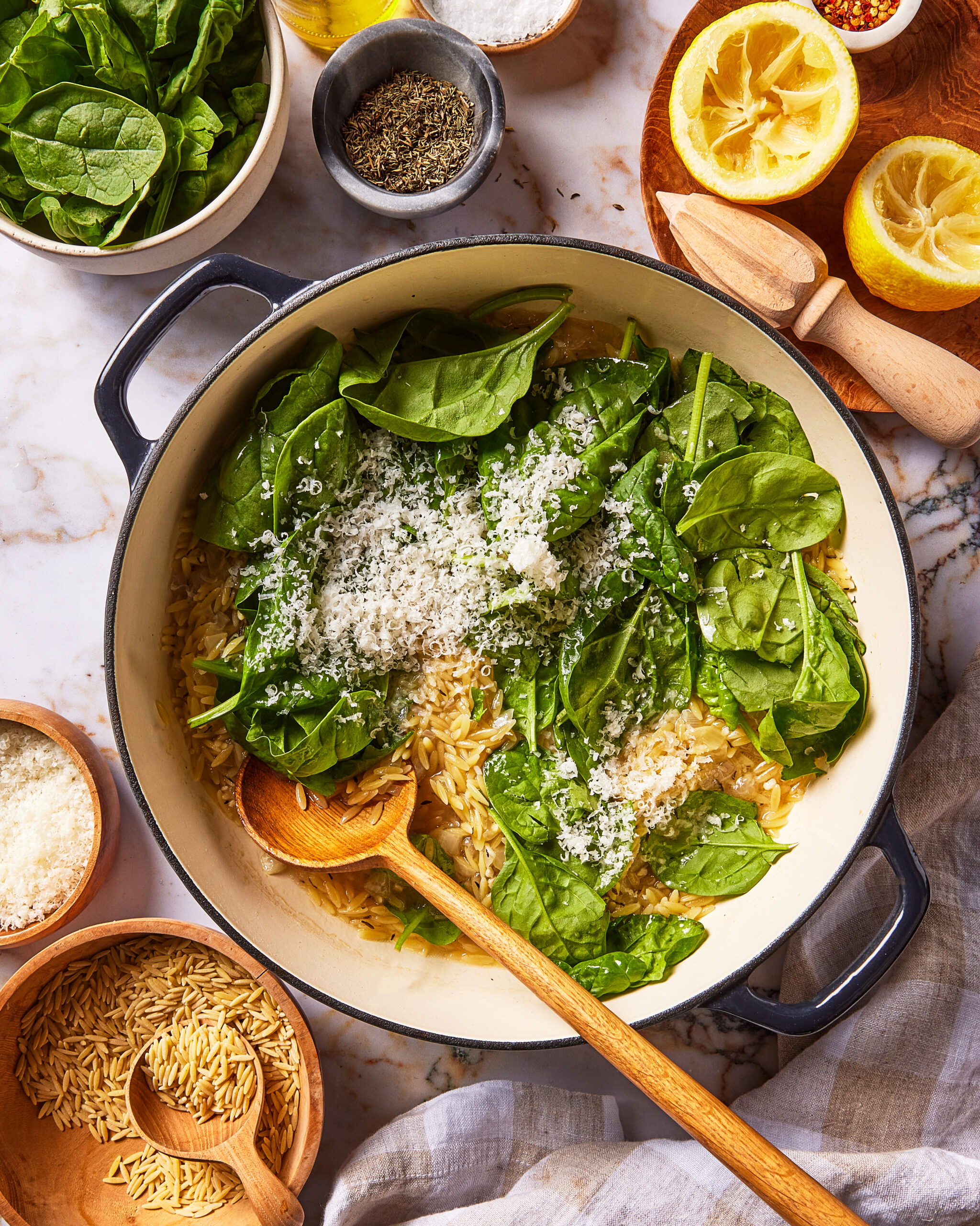 A large pan of orzo being mixed with fresh spinach and grated cheese, surrounded by bowls of dry orzo, spinach, and halved lemons on a marble surface.