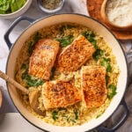 A pan of creamy orzo with spinach and four seared salmon fillets, served with bowls of spinach, herbs, chili flakes, and grated cheese.