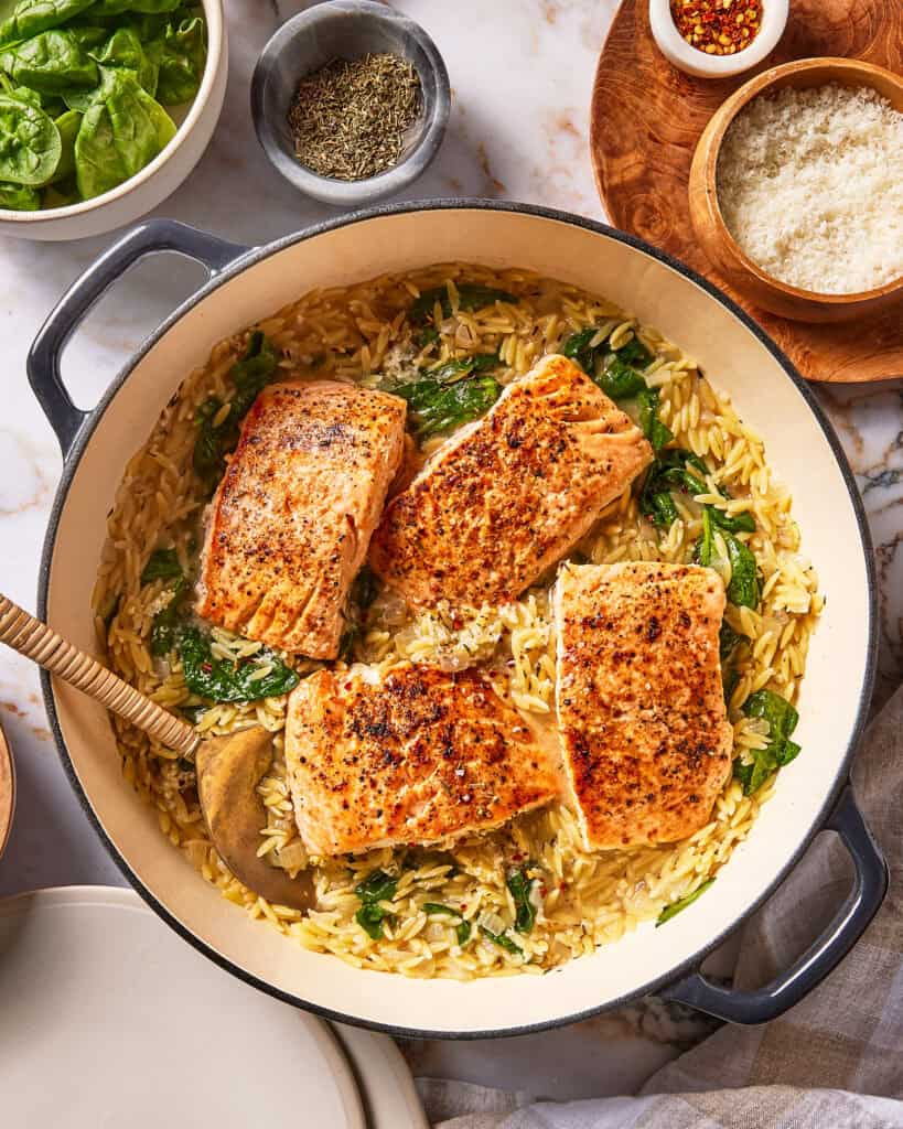 A pan of creamy orzo with spinach and four seared salmon fillets, served with bowls of spinach, herbs, chili flakes, and grated cheese.