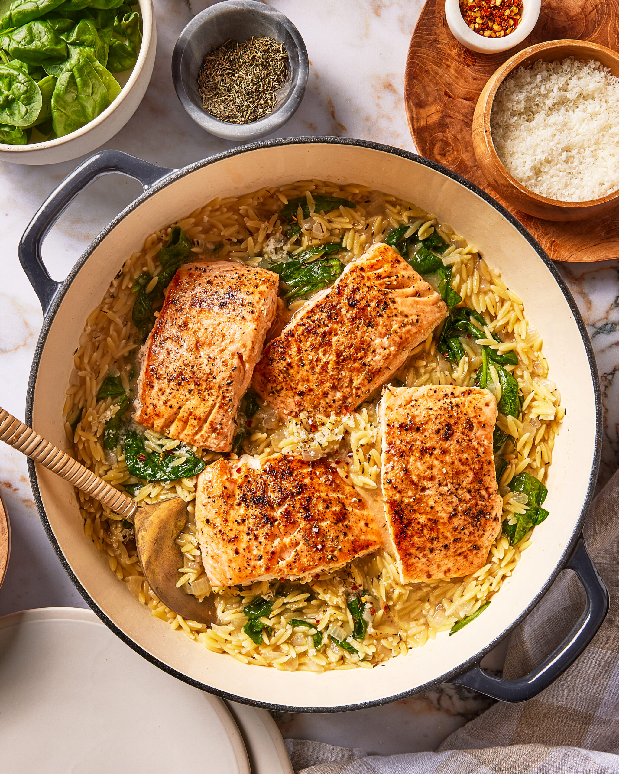 A pan of creamy orzo with spinach and four seared salmon fillets, served with bowls of spinach, herbs, chili flakes, and grated cheese.