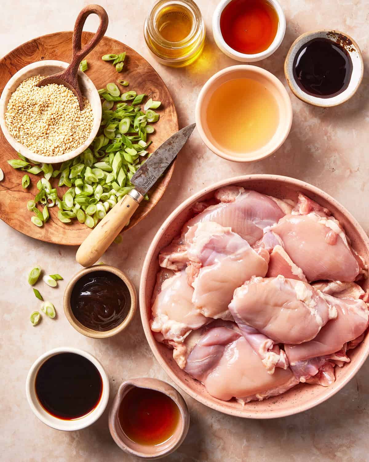 A large bowl of raw chicken thighs surrounded by small bowls of sesame seeds, sliced green onions, and various liquid seasonings.