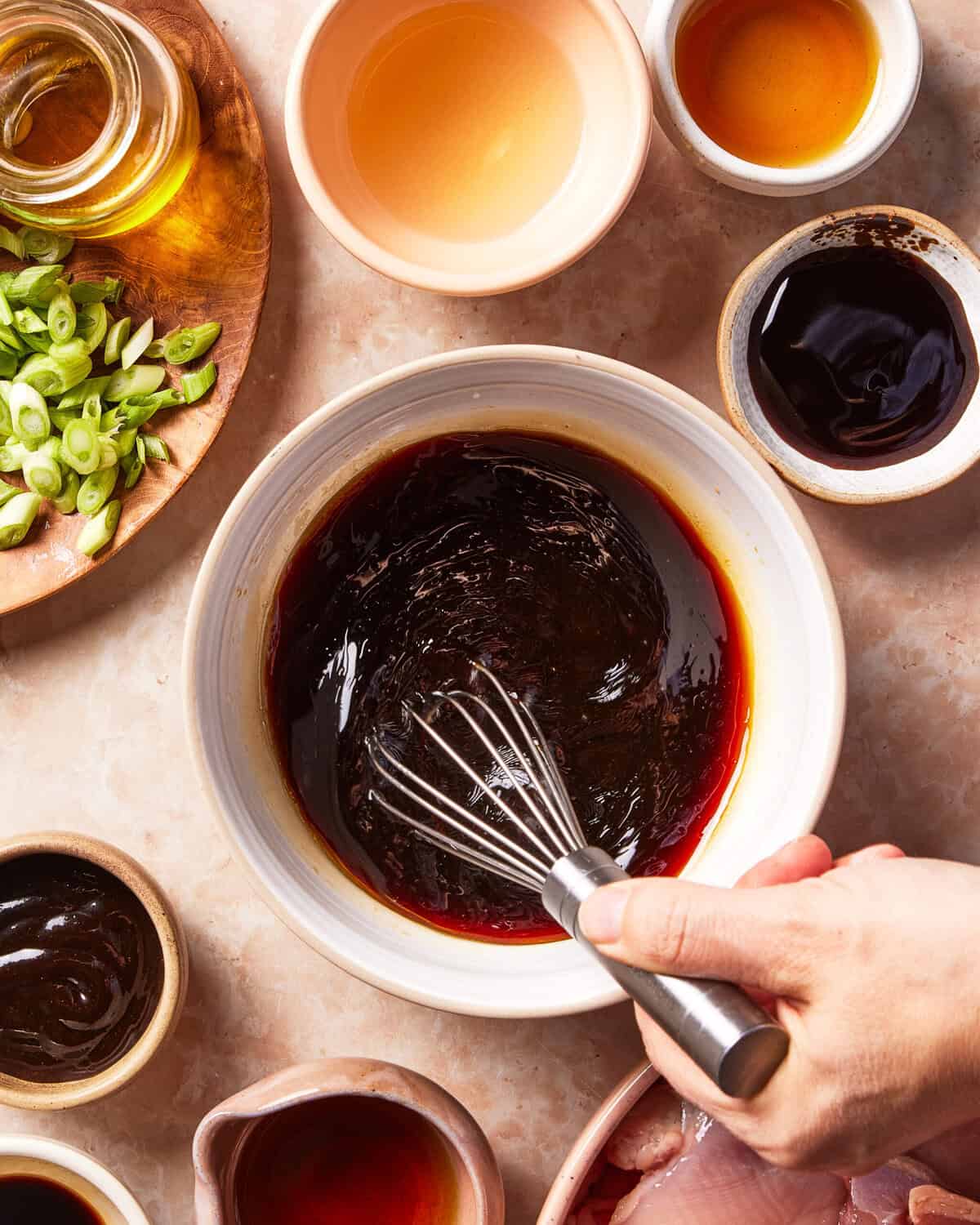 A hand whisks a dark sauce in a bowl, surrounded by small dishes of oil, scallions, and other liquid ingredients.