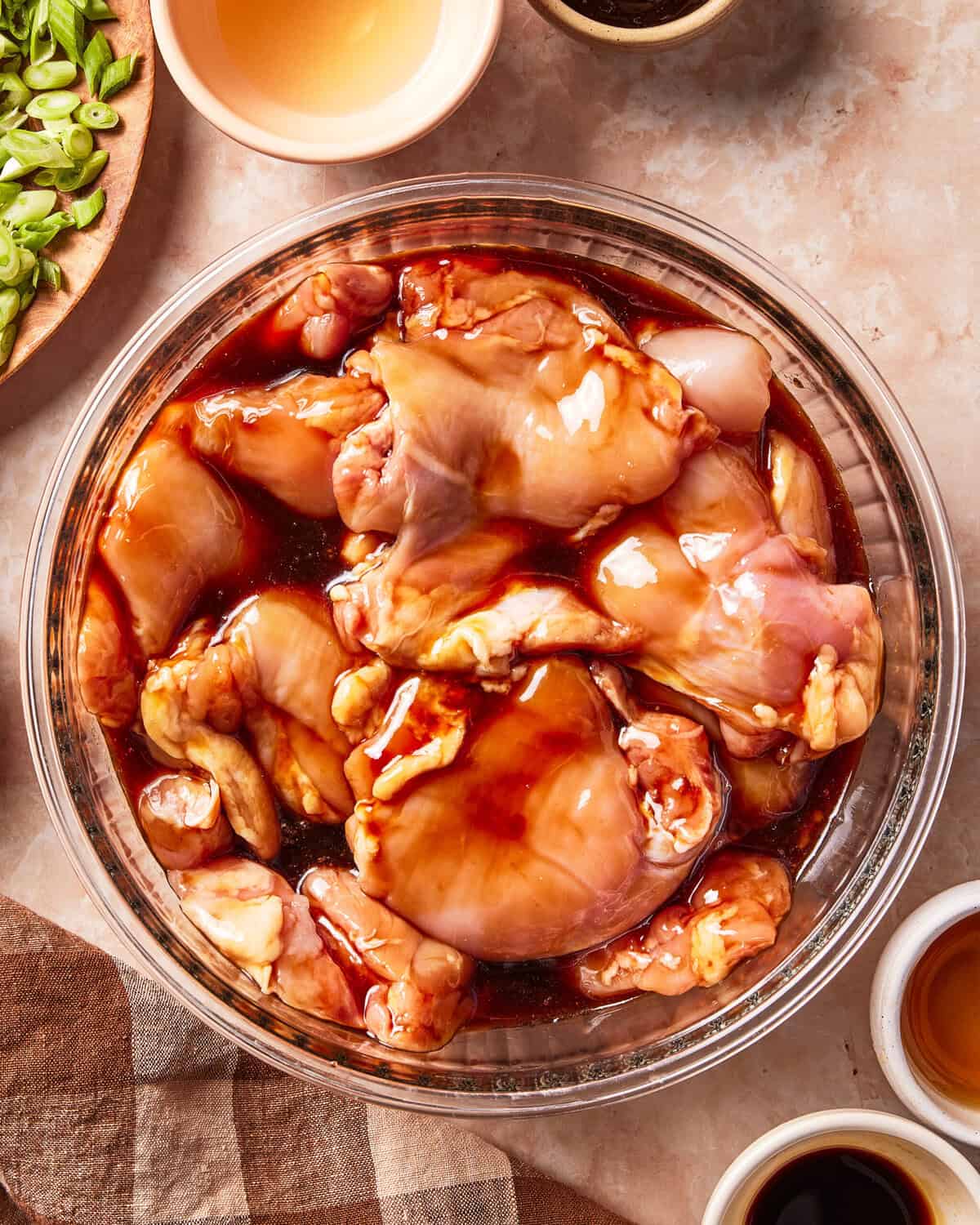 Raw chicken pieces marinating in a dark soy-based sauce inside a glass bowl, with small bowls of sauce and chopped scallions nearby.