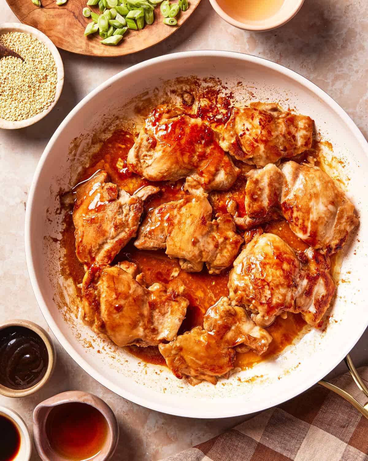 Glazed chicken thighs cooking in a large white skillet, surrounded by small bowls of sesame seeds, scallions, and sauces.