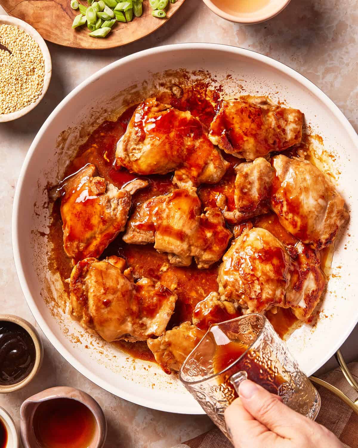 A hand pours sauce into a skillet filled with cooked chicken thighs, surrounded by small bowls of sesame seeds, scallions, and sauce.