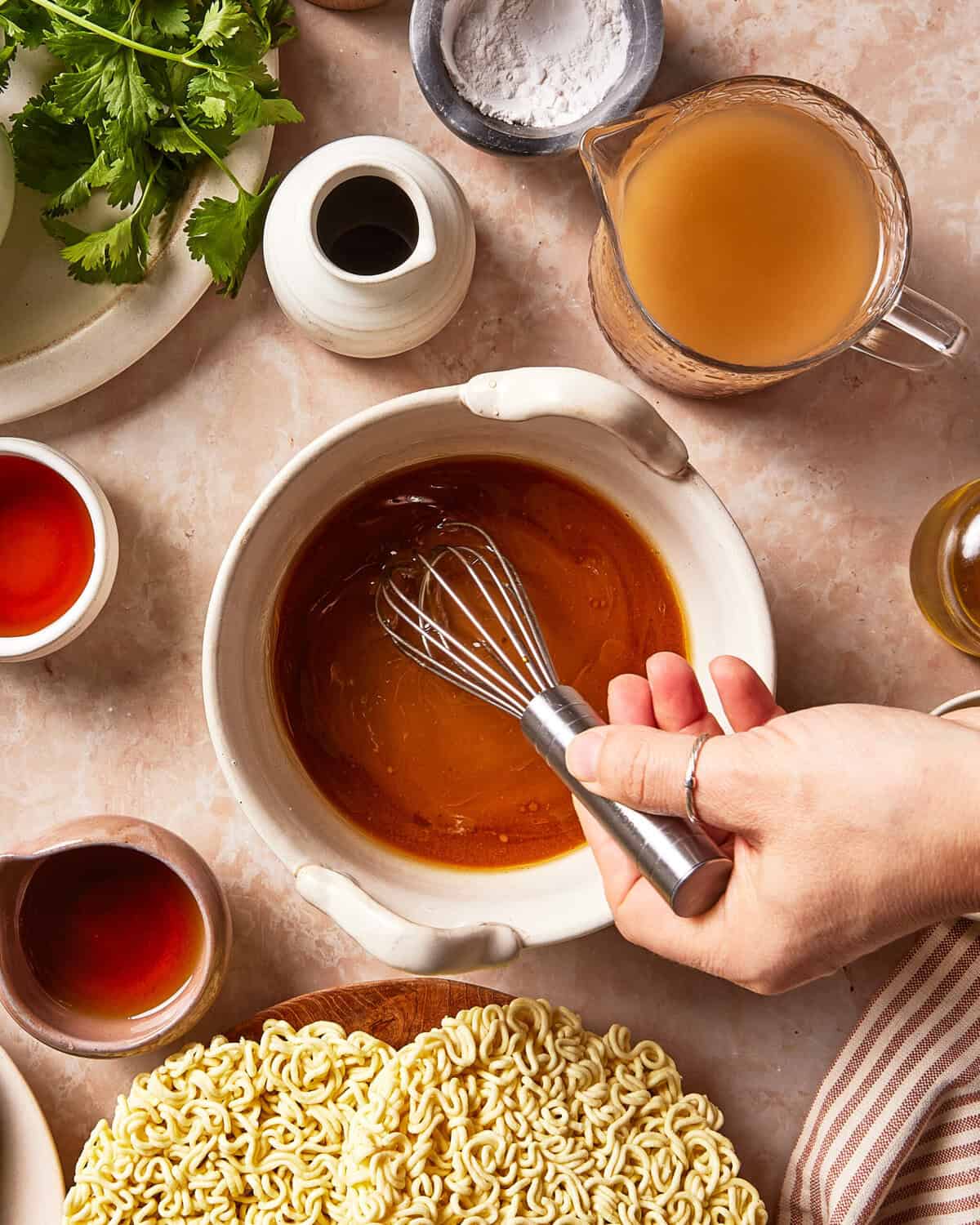 A hand uses a wire whisk to mix a brown sauce in a ceramic bowl, surrounded by ingredients including ramen noodles, cilantro, broth, and various sauces.