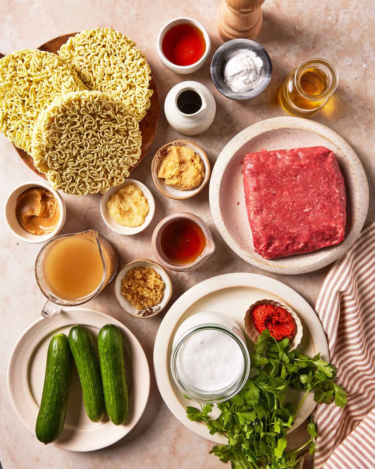Ingredients for ramen, including dry noodles, ground meat, broth, coconut milk, peanut butter, cucumbers, fresh cilantro, chili paste, and various sauces in small bowls.