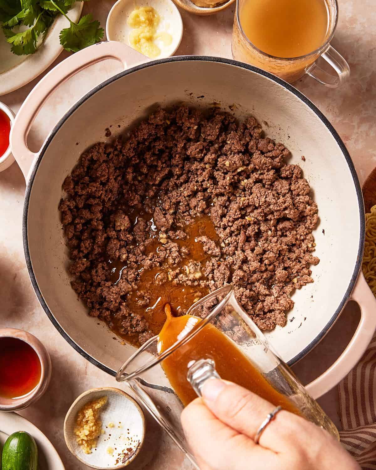 A hand pours broth from a glass measuring cup into a Dutch oven filled with browned ground meat.