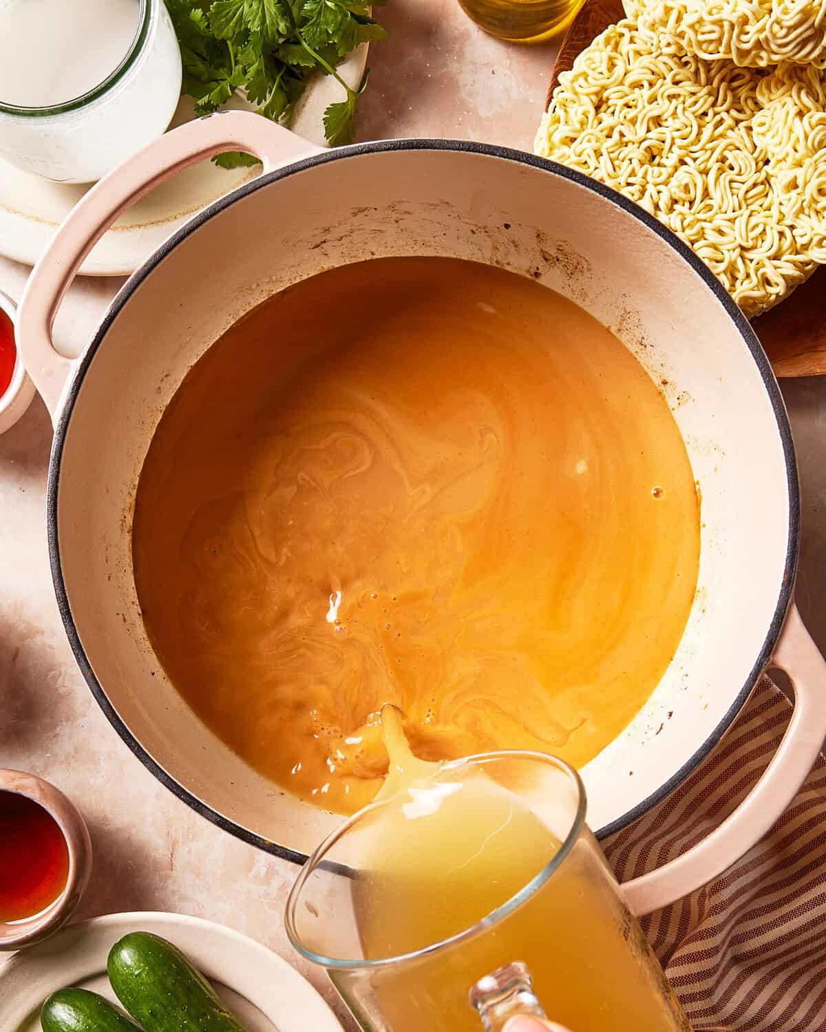 A hand pours broth from a glass pitcher into a large pot of orange soup, surrounded by ingredients including dried ramen noodles, cilantro, and cucumbers.