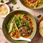Two bowls of spicy ramen topped with ground meat, fresh cucumber slices, cilantro, and sesame seeds, with a ceramic spoon in the foreground.
