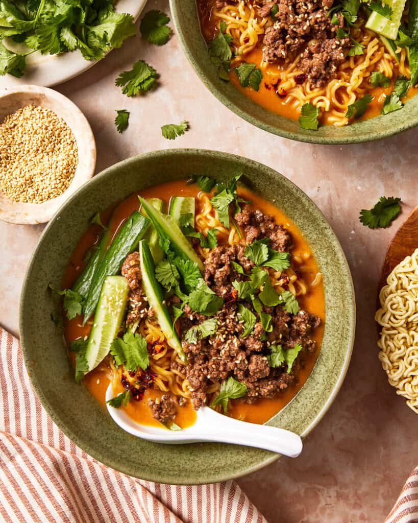 Two bowls of spicy ramen topped with ground meat, fresh cucumber slices, cilantro, and sesame seeds, with a ceramic spoon in the foreground.