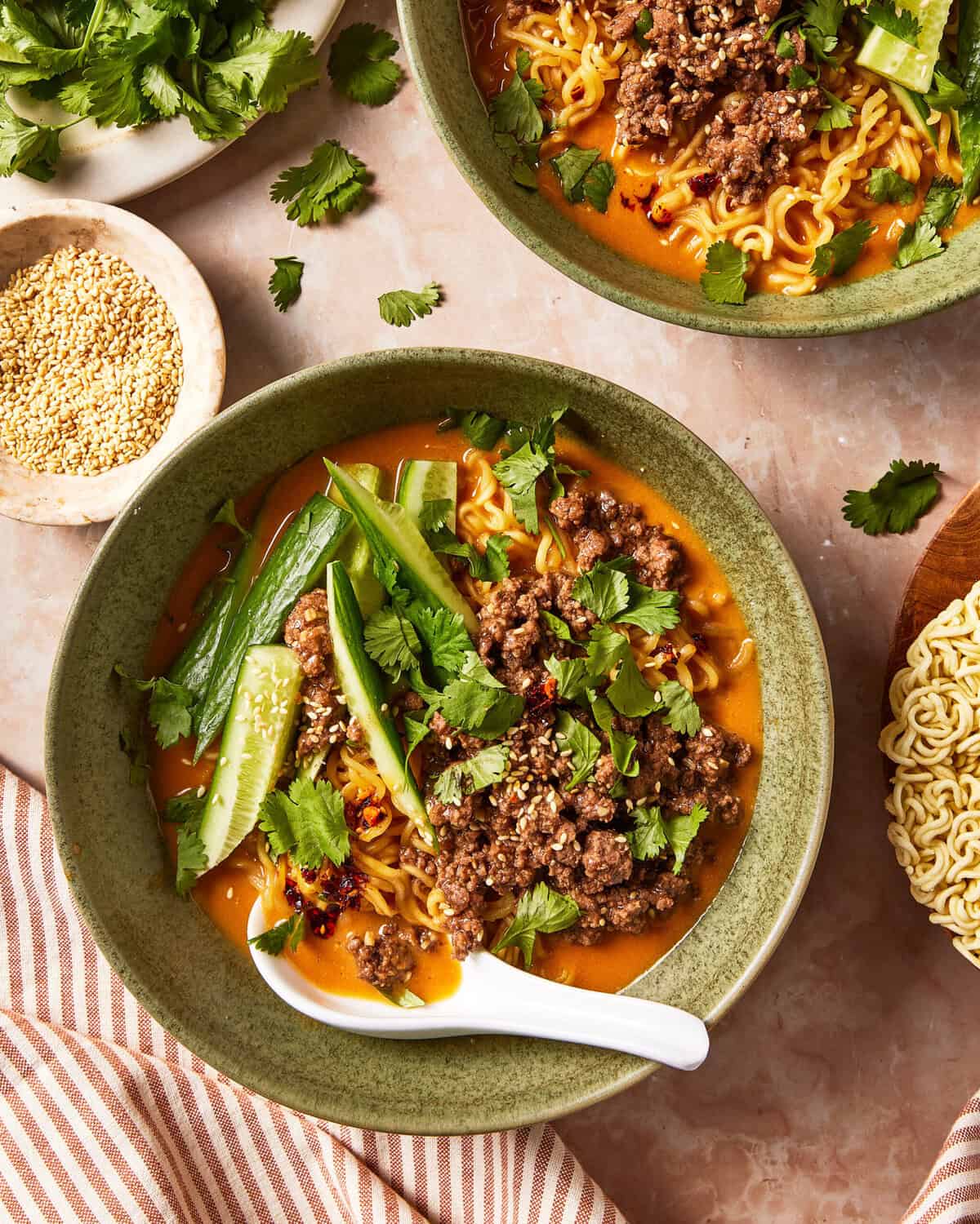 Two bowls of spicy ramen topped with ground meat, fresh cucumber slices, cilantro, and sesame seeds, with a ceramic spoon in the foreground.