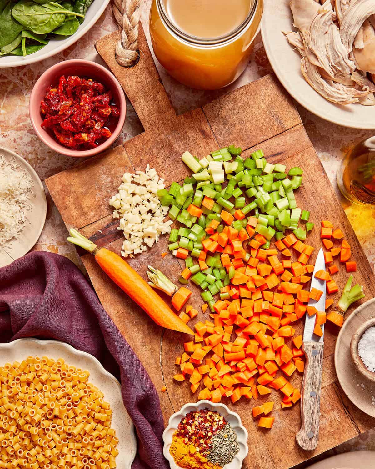 A wooden cutting board with diced carrots and celery, a pile of minced garlic, and a whole carrot, surrounded by bowls of pasta, shredded chicken, sun-dried tomatoes, broth, and seasonings.