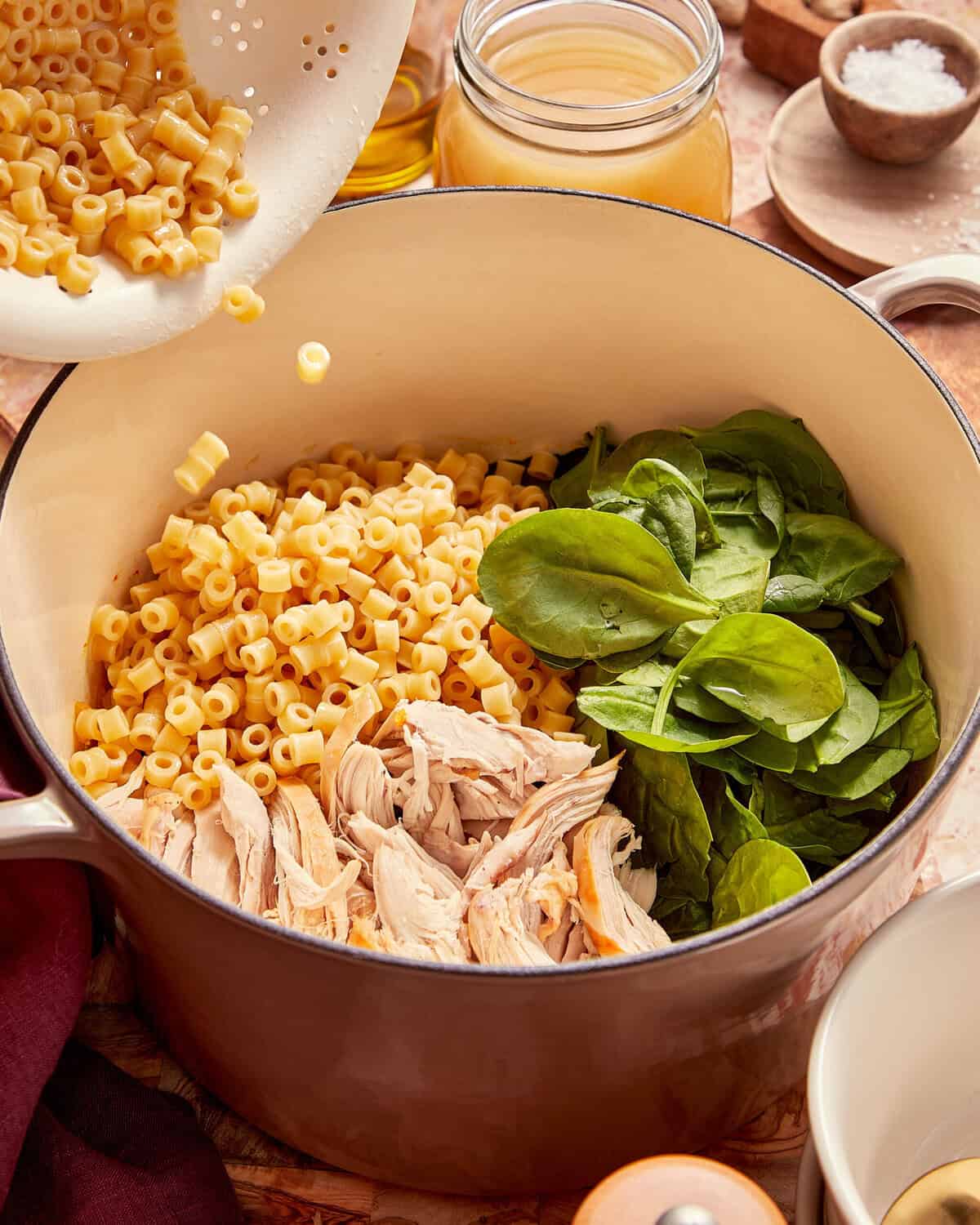 A colander pours ditalini pasta into a large pot containing shredded chicken and fresh spinach.