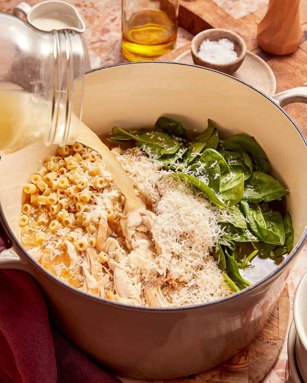 Liquid being poured from a glass jar into a Dutch oven filled with shredded chicken, pasta, spinach, and shredded cheese.