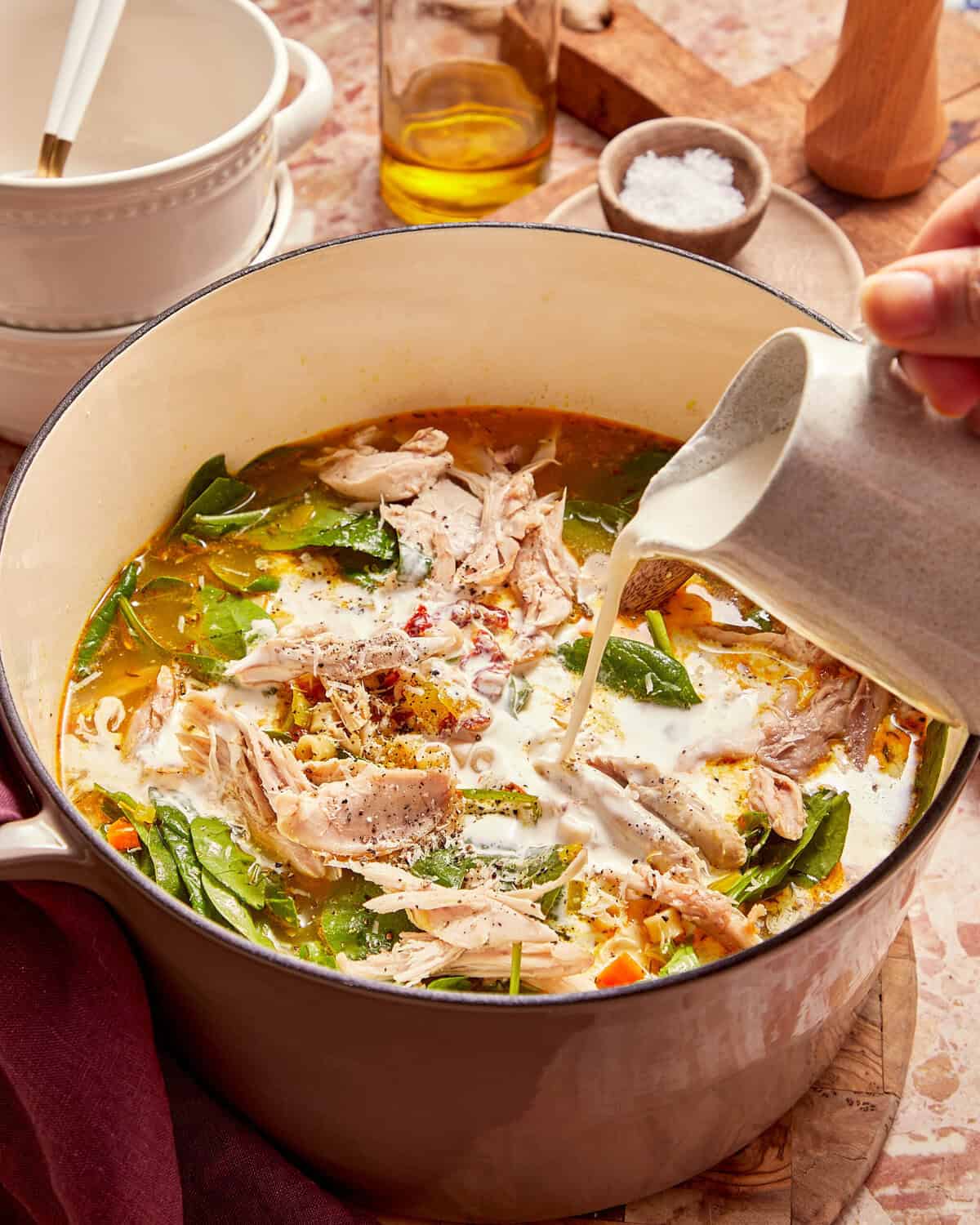 Cream being poured from a small pitcher into a pot of chicken and spinach soup.