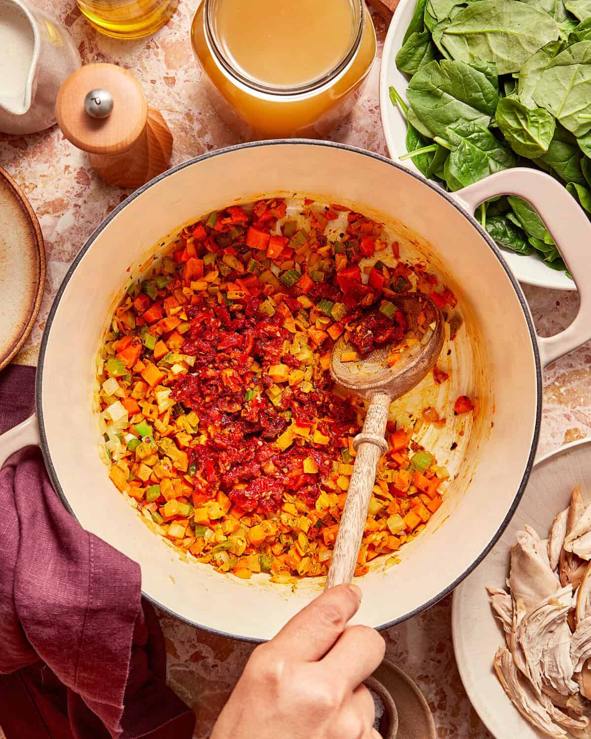 A hand stirs sautéed, diced vegetables and red tomato paste in a large enameled pot, surrounded by bowls of spinach and shredded chicken.