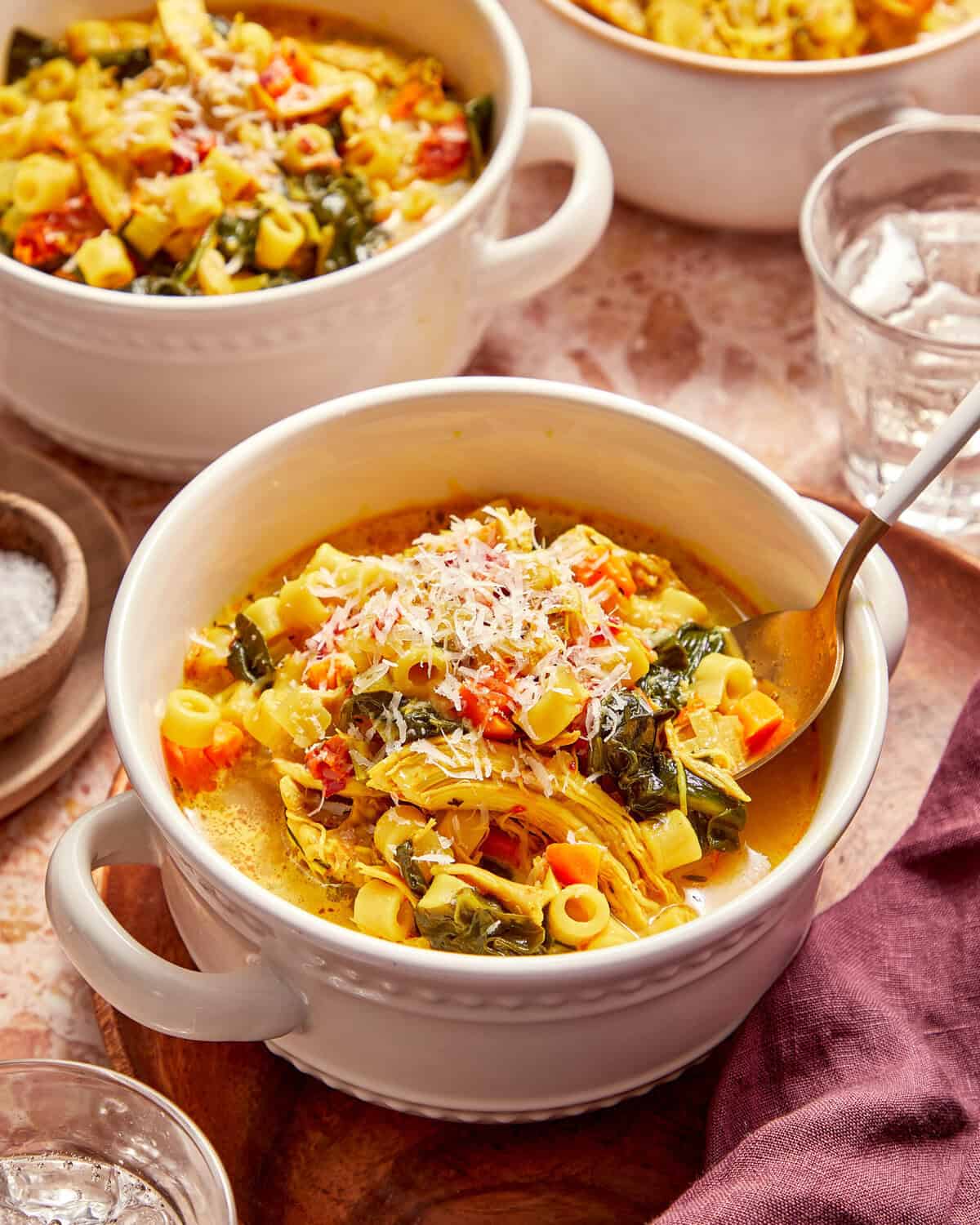 A bowl of chicken noodle soup with leafy greens, topped with grated cheese and served with a spoon.