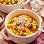 A bowl of chicken and pasta soup with greens, topped with grated cheese and cracked pepper.