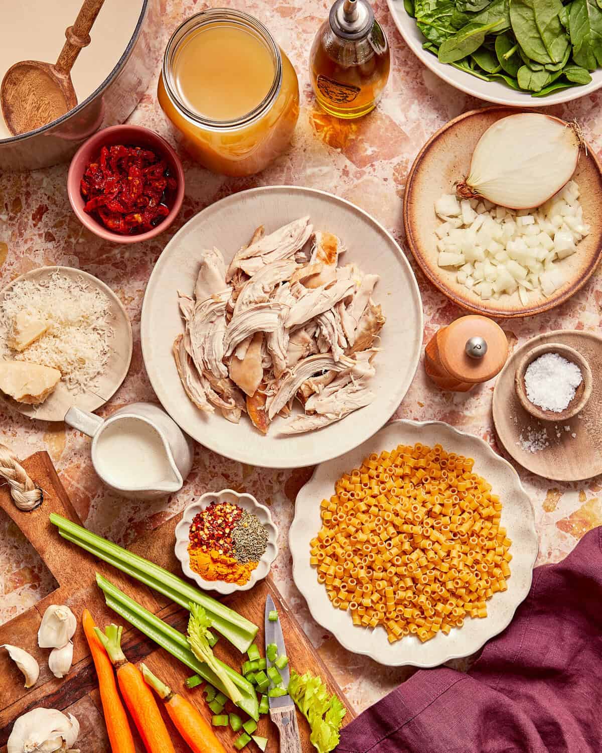 Overhead view of ingredients for chicken soup: shredded chicken, ditalini pasta, onion, carrots, celery, broth, spices, cream, and spinach on a marble countertop.