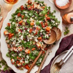 A large platter of roasted sweet potato and kale salad with feta, dried cranberries, and grains, served with wooden salad spoons.