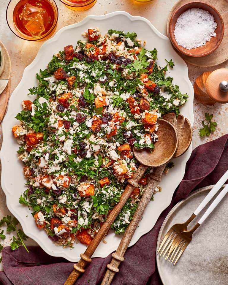 A large platter of roasted sweet potato and kale salad with feta, dried cranberries, and grains, served with wooden salad spoons.
