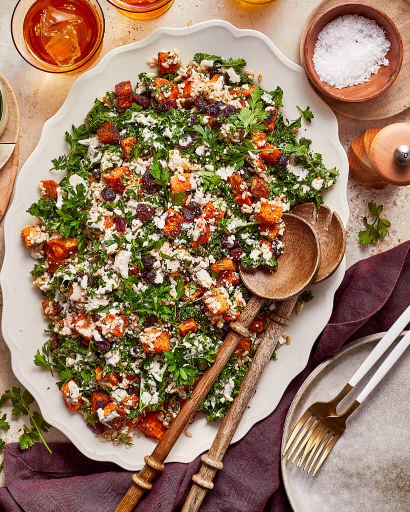 A large platter of roasted sweet potato and kale salad with feta, dried cranberries, and grains, served with wooden salad spoons.