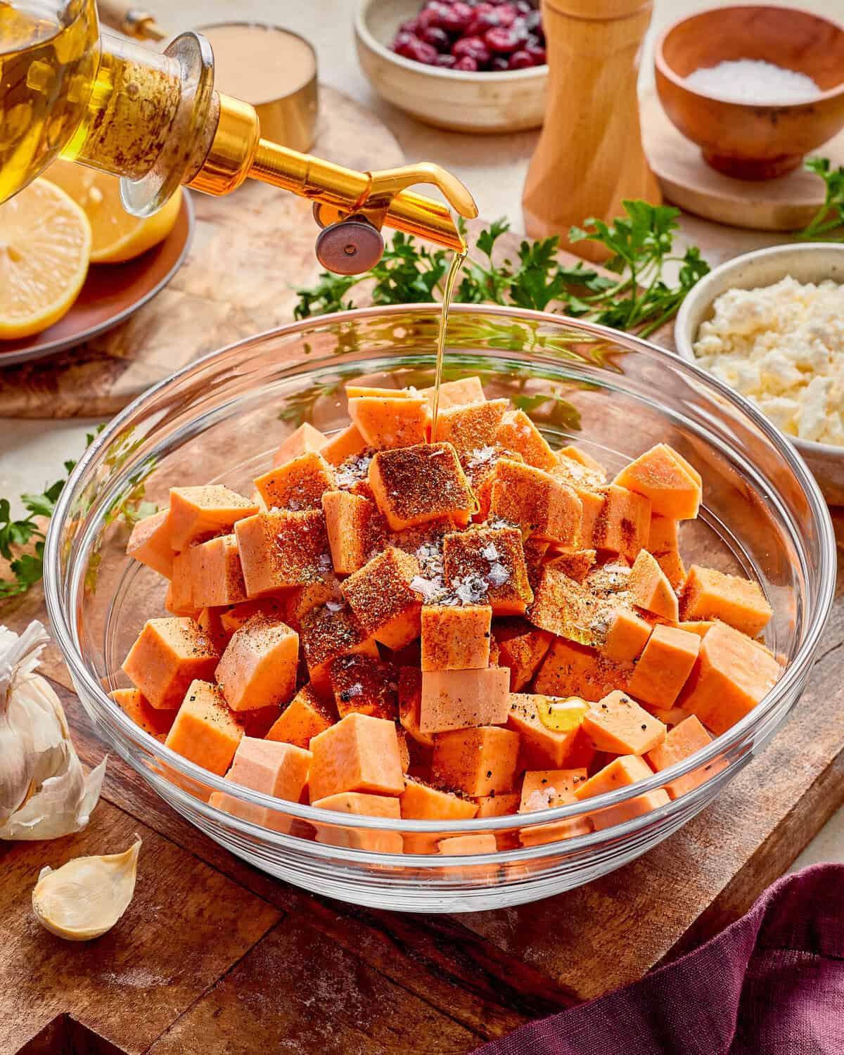 Olive oil being poured from a glass cruet onto a glass bowl of cubed sweet potatoes seasoned with spices.