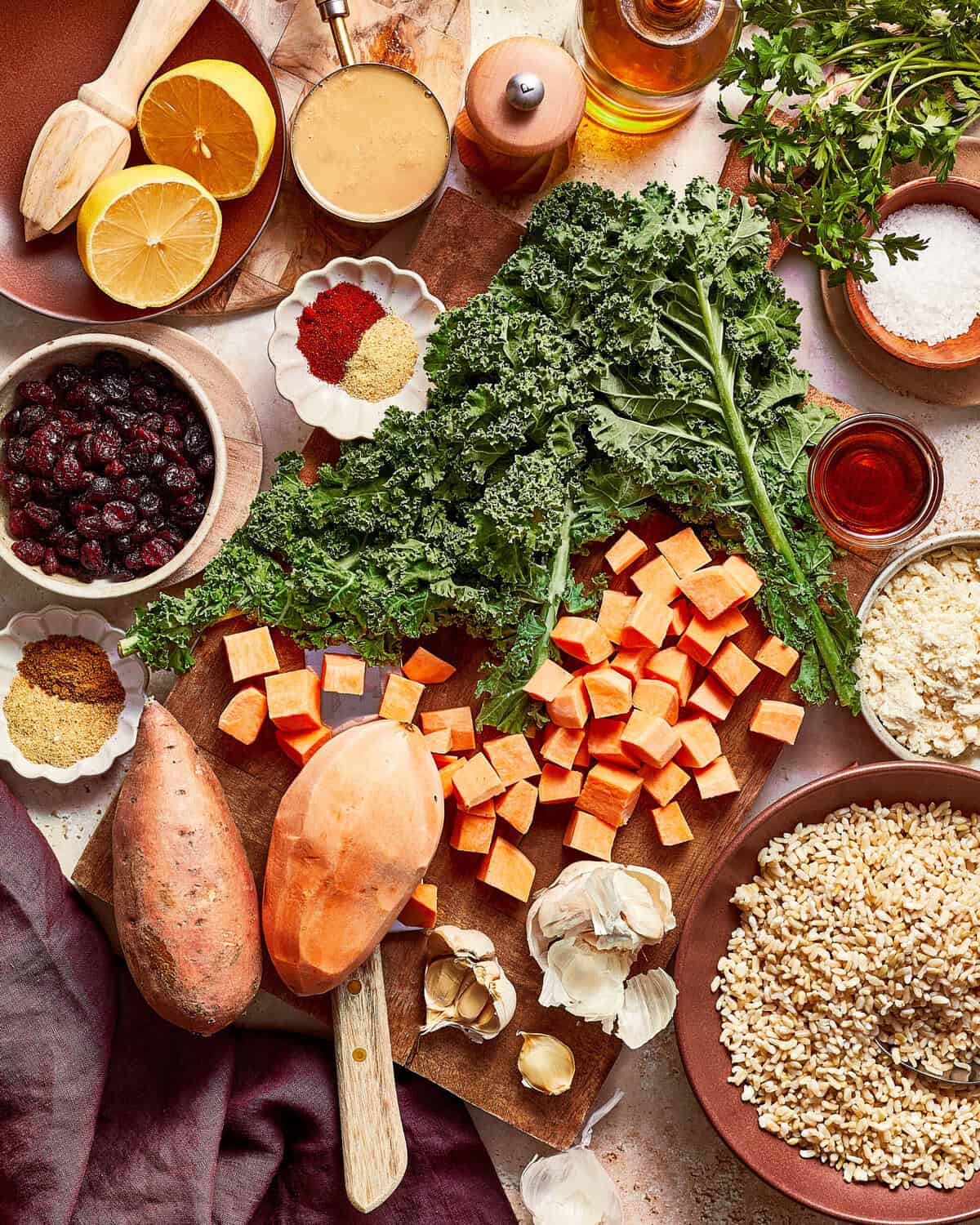An overhead view of raw ingredients for a salad, including curly kale, cubed sweet potatoes, dried cranberries, brown rice, garlic, lemon, and spices arranged on a wooden board.