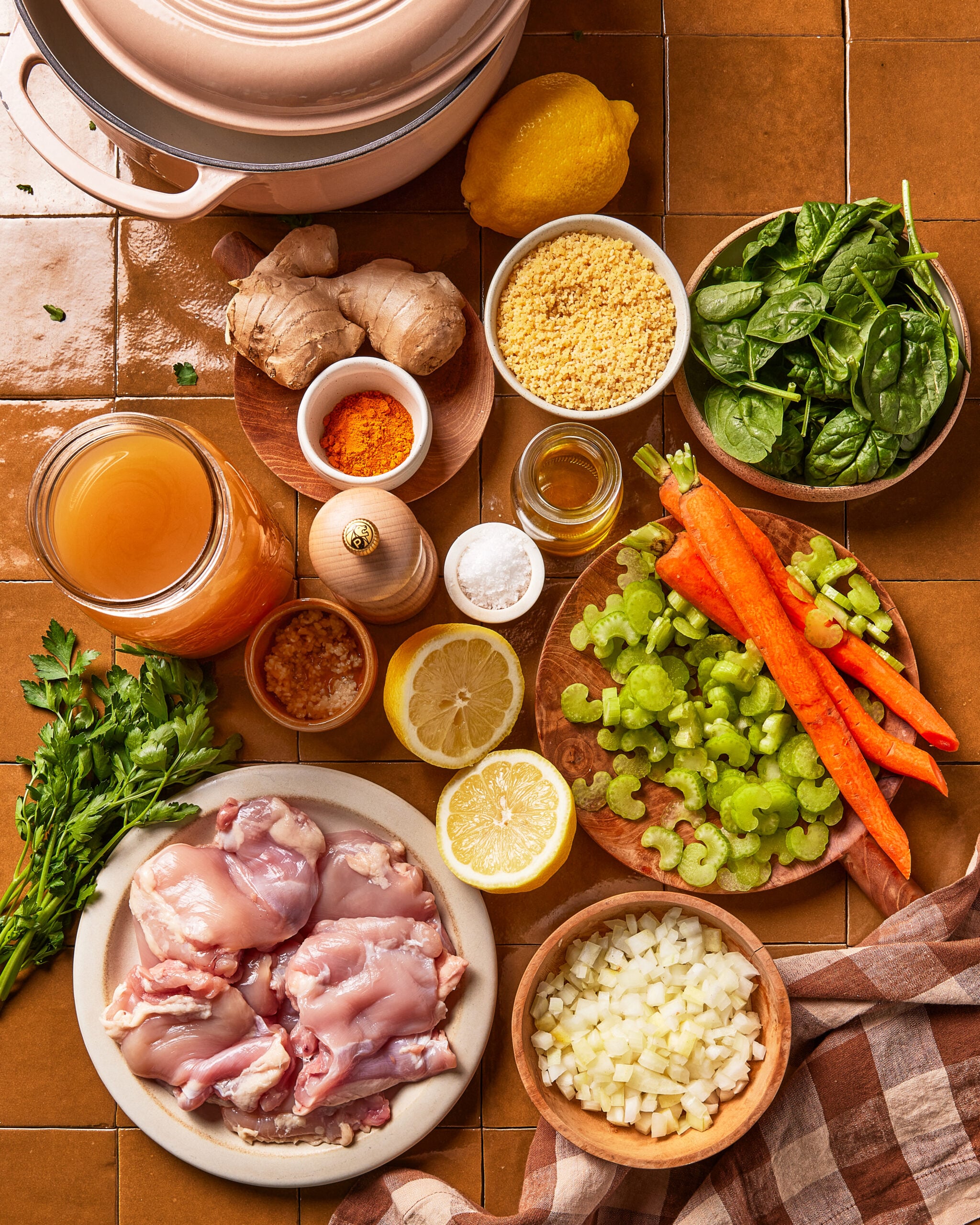 Top-down view of raw chicken, chopped onions, carrots, celery, spinach, ginger, parsley, lemon, stock, spices, garlic, and couscous on a tiled countertop, ready for cooking.