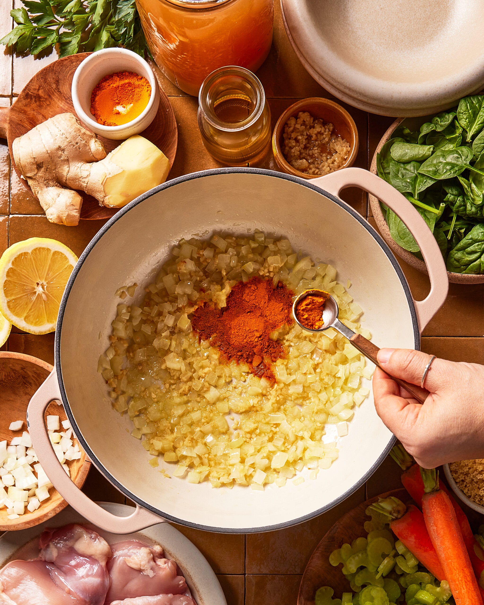 A hand adds paprika to sautéed onions in a white pot. Surrounding the pot are ingredients like raw chicken, lemon, ginger, garlic, spinach, carrot, celery, spices, and broth.