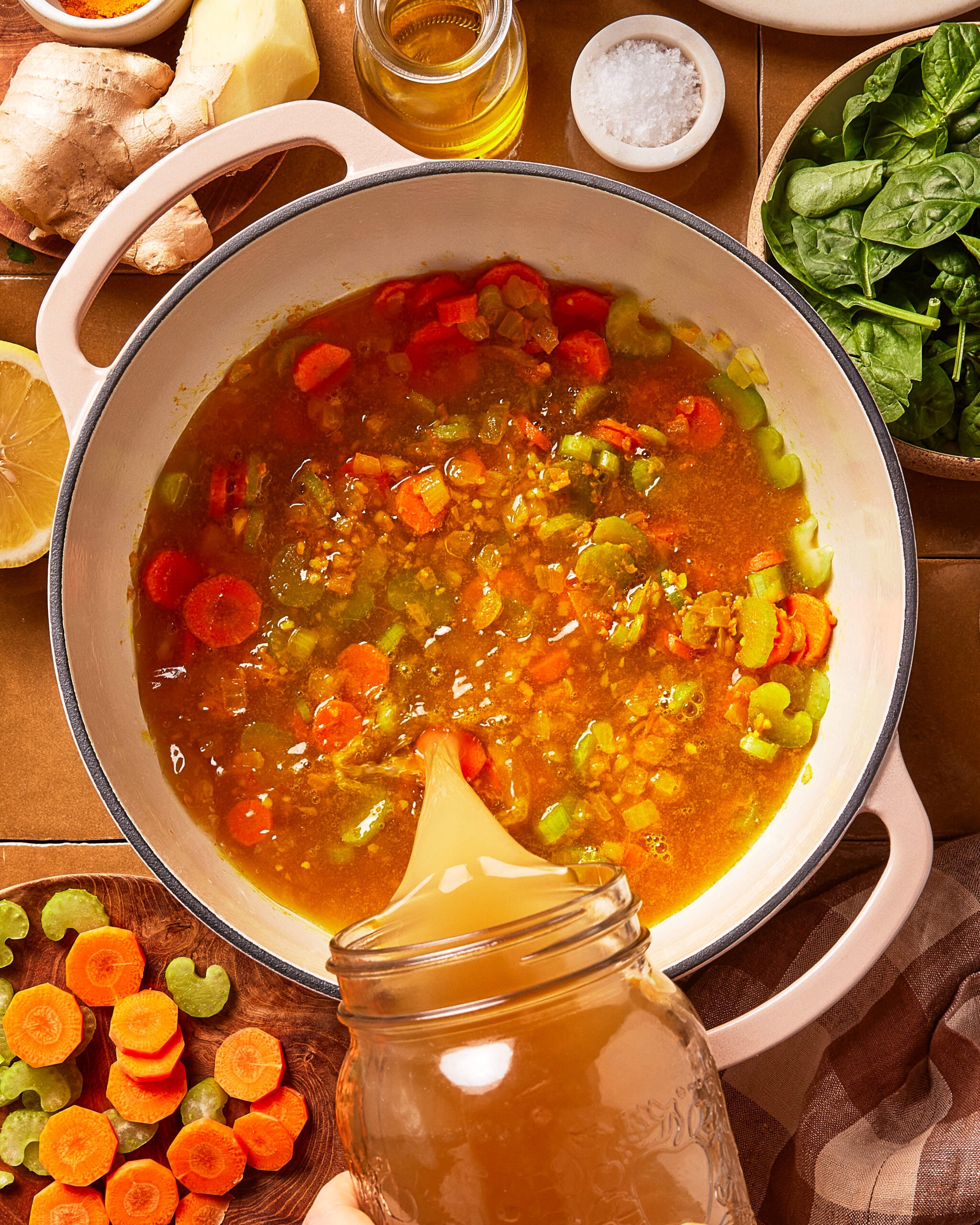 A pot of soup with chopped carrots, celery, and broth being poured in from a jar. Fresh spinach, ginger, lemon, and seasonings are arranged around the pot.